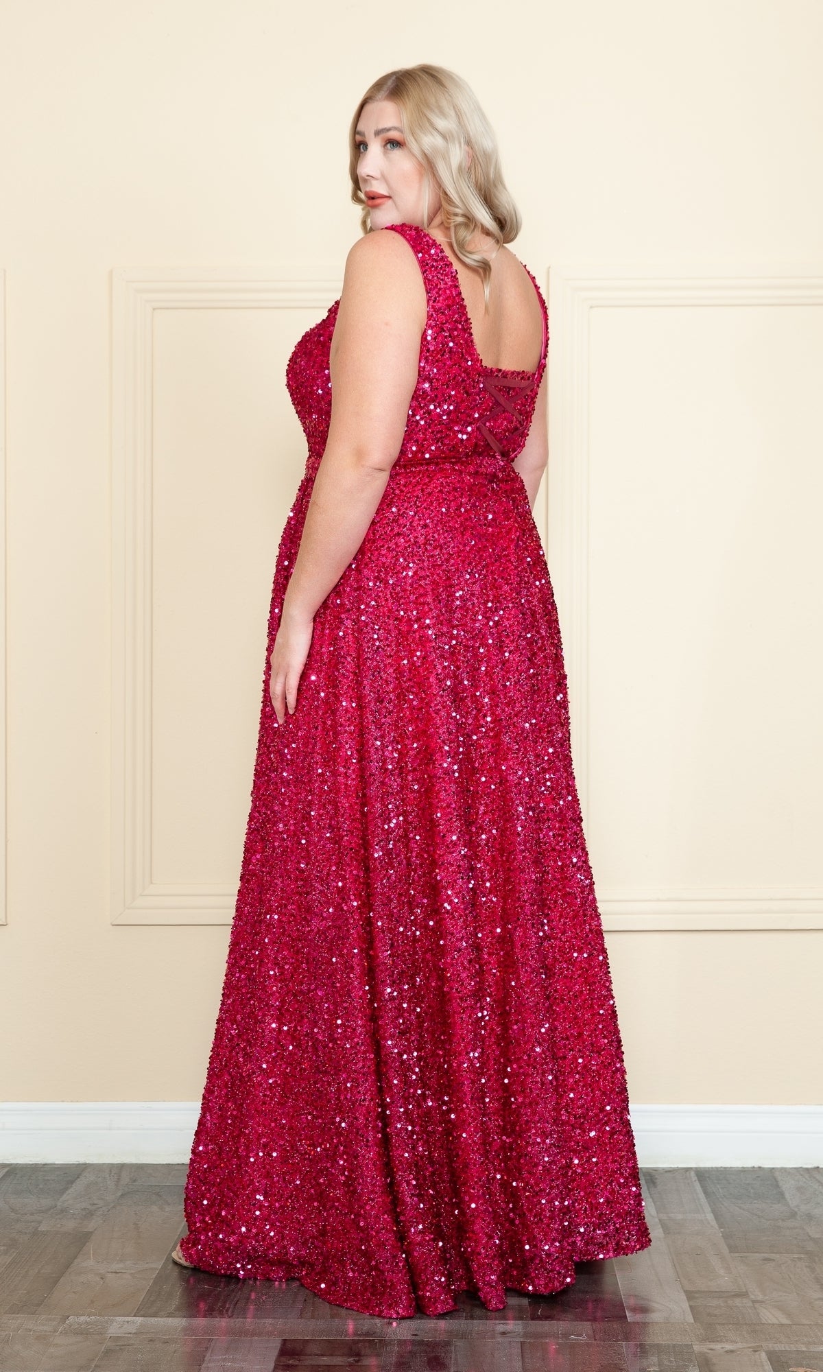 A woman in the Plus Long Sequin Prom Dress with Underskirt W1128, featuring a sleeveless floor-length design and corset lace-up back, stands on a wooden floor facing slightly to the side against a cream-colored wall.